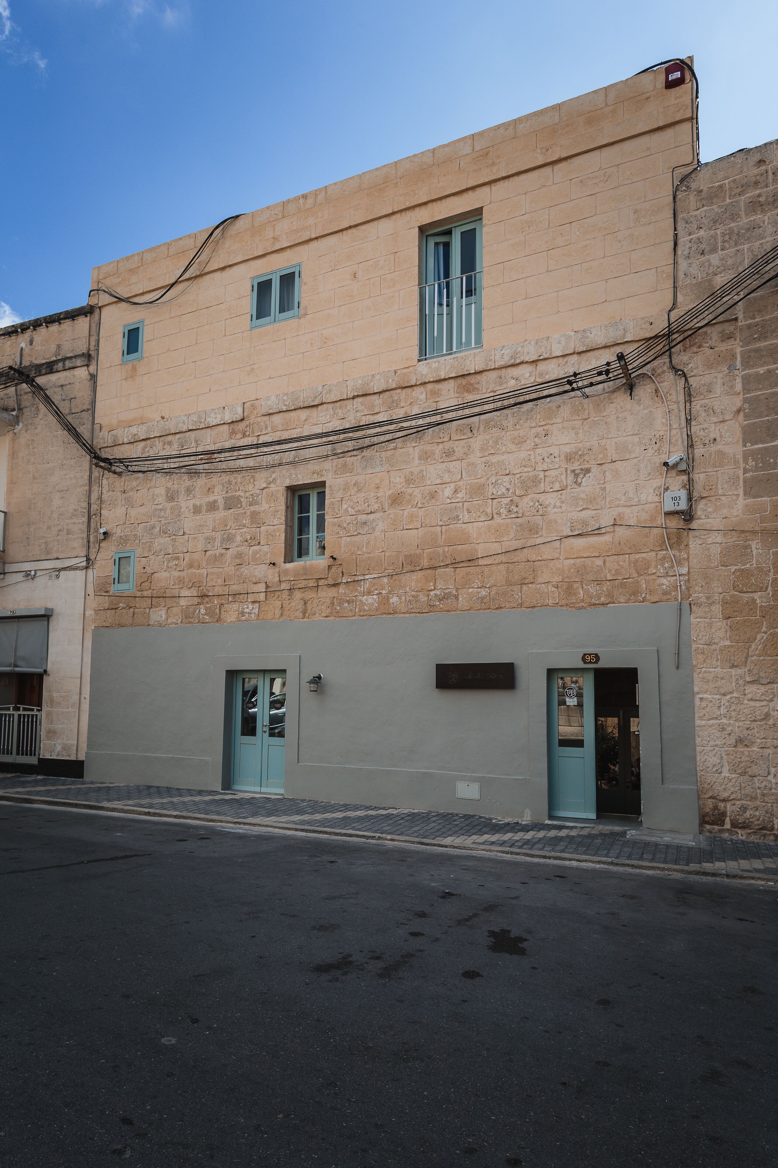 Un edificio de dos pisos con fachada de piedra color beige, una puerta azul y un ventanal en la planta baja.