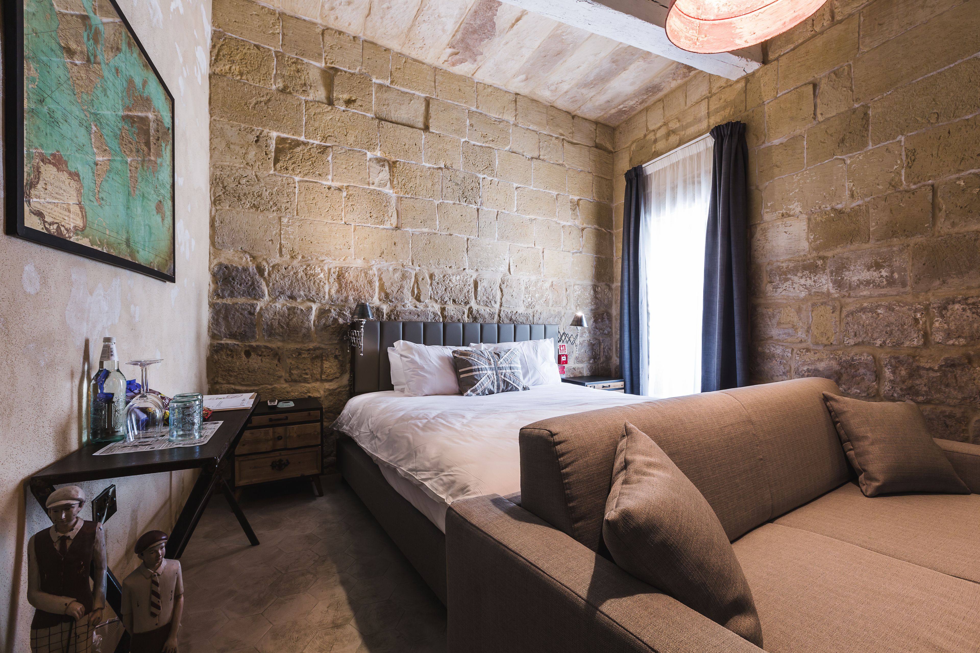 A bedroom with a brick wall, a bed with white linens, a brown sofa, and a small table with items on it.