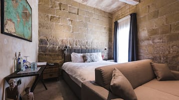 A bedroom with a brick wall, a bed with white linens, a brown sofa, and a small table with items on it.