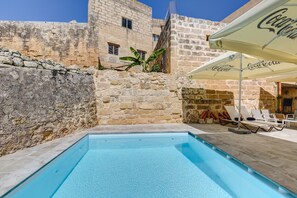 A swimming pool with lounge chairs and umbrellas, set against a stone wall and a building with windows. - Talbot and Bons Boutique Bed & Breakfast (Gudja)