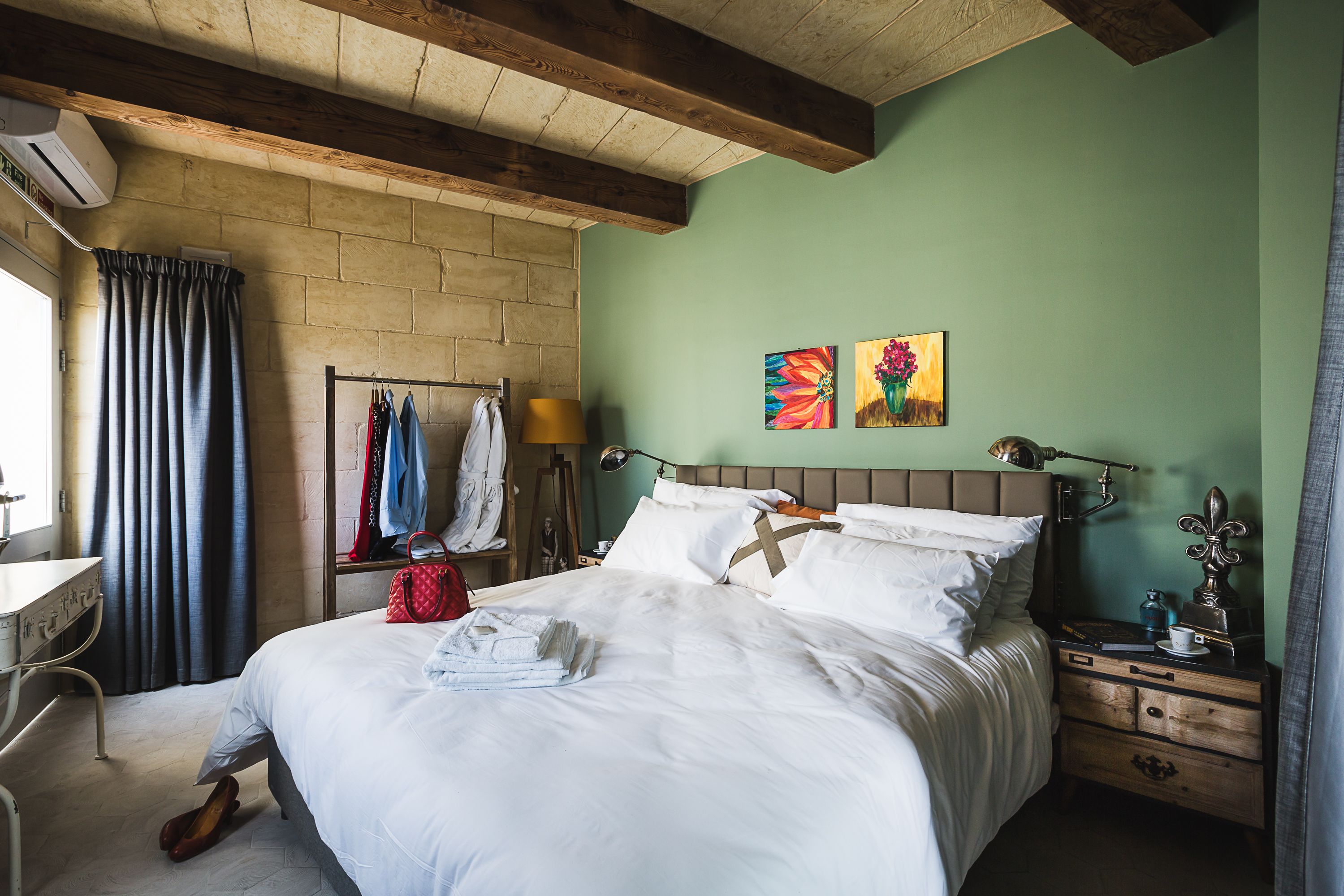 Uma cama bem arrumada com roupa de cama branca, uma mesa de cabeceira de madeira com abajur e uma parede com duas pinturas coloridas.