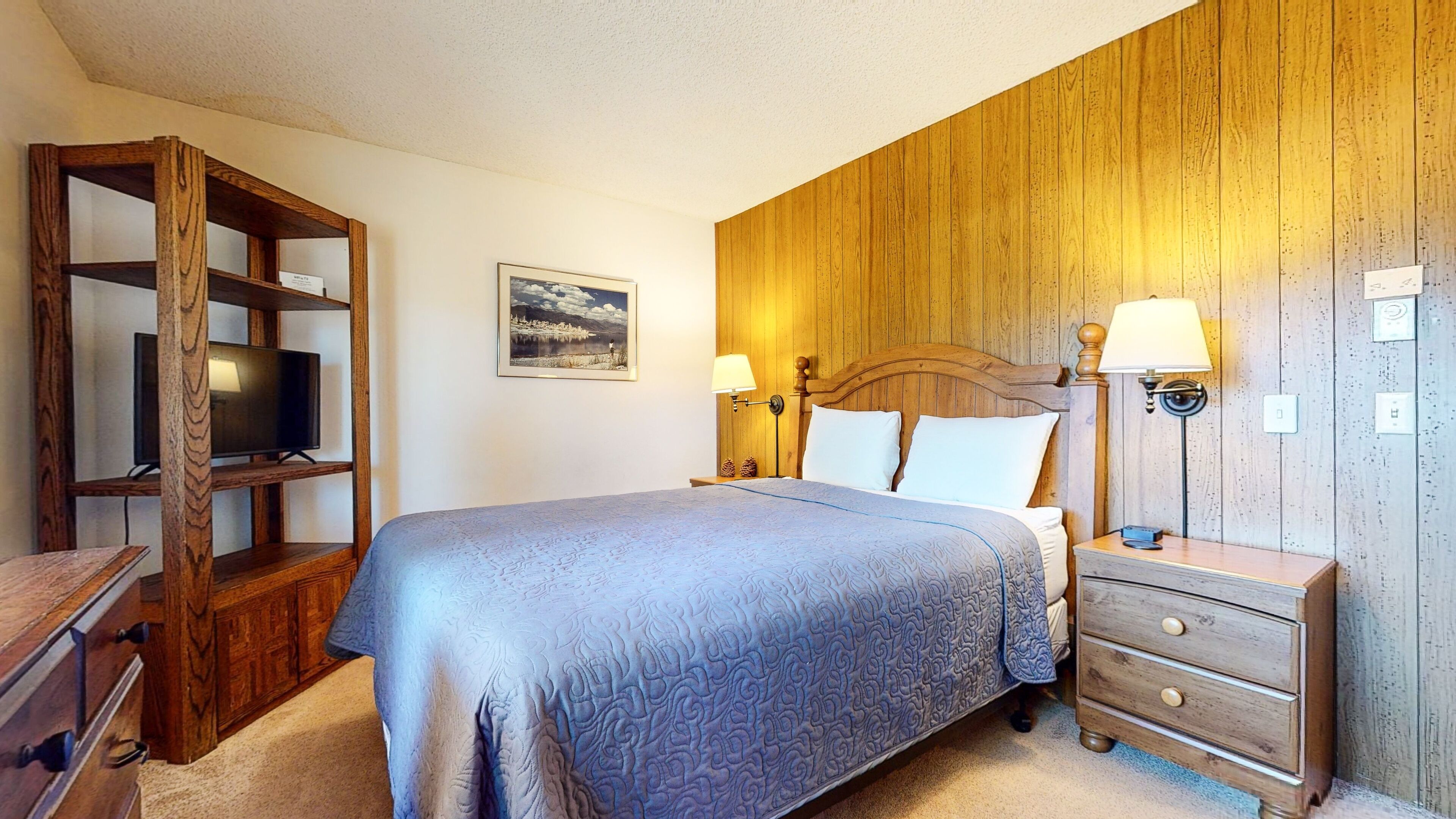 A bedroom with a wooden headboard, a bed with a blue patterned bedspread, two bedside lamps, a wooden nightstand, a television on a wooden shelf, and a framed picture on the wall.