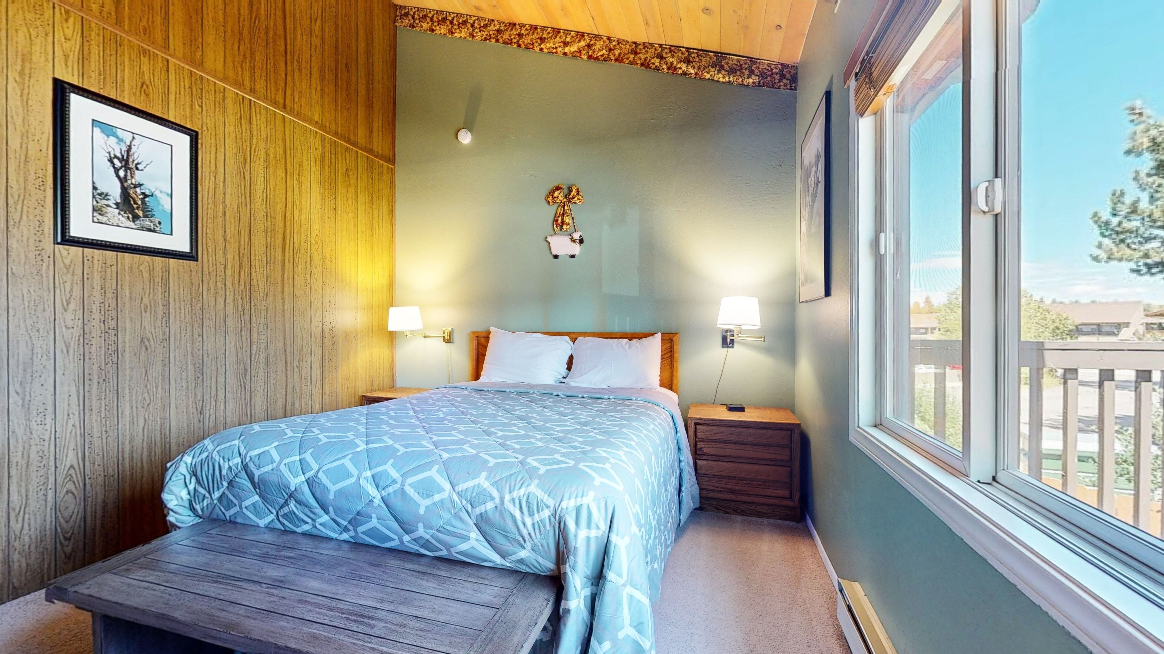 A bedroom with a wooden headboard, a bed with a blue patterned bedspread, two bedside lamps, a small wooden nightstand, and a window with a view of the outside.