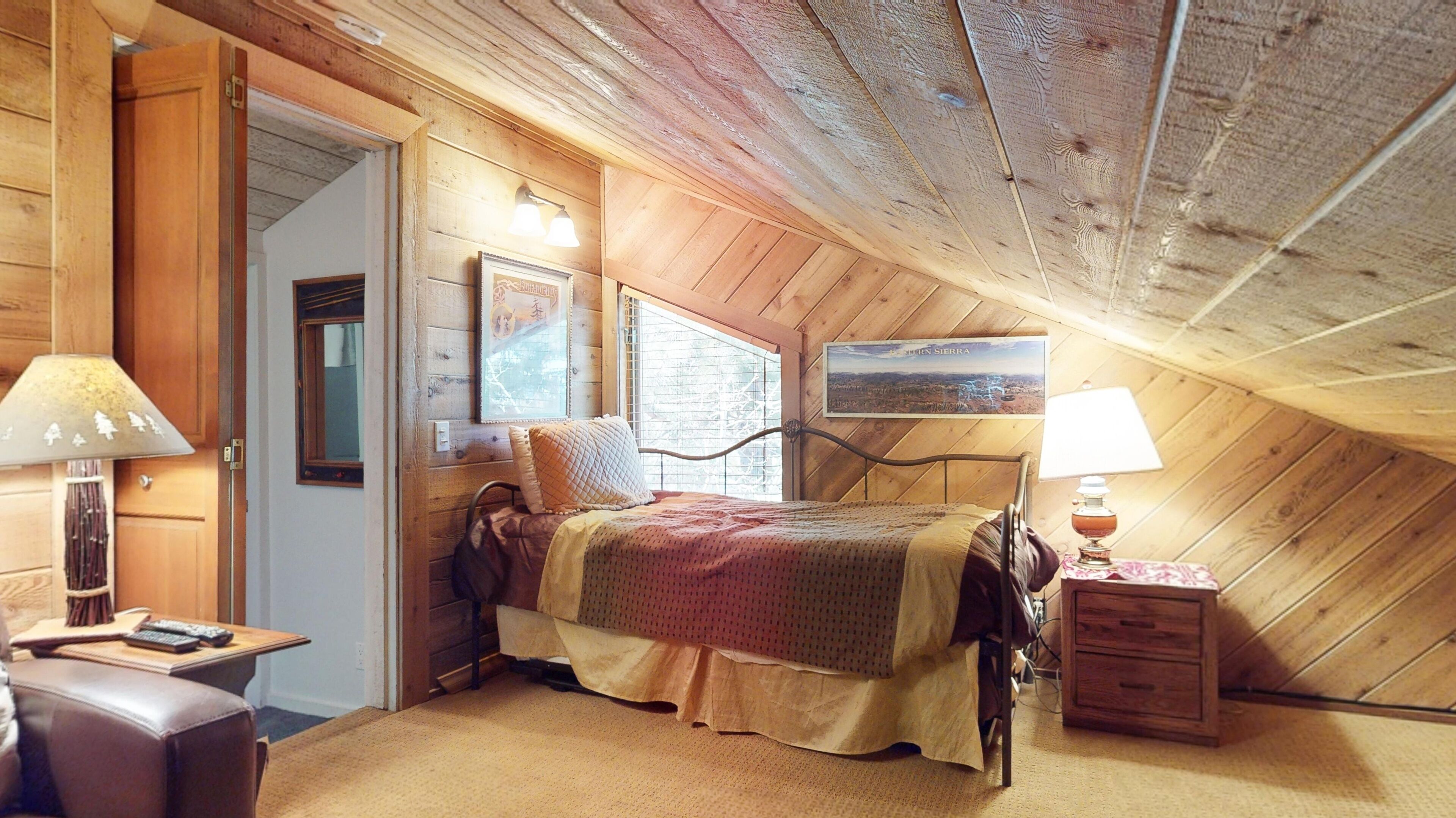A cozy attic bedroom with a wooden ceiling, a bed with a quilt, a nightstand with a lamp, and a window with blinds.