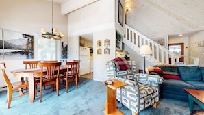 A spacious living room with a high ceiling, a dining area with wooden furniture, and a staircase leading to an upper level.
