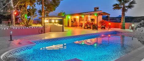 A swimming pool with a tiled edge, a covered outdoor seating area, and a house with a patio.