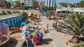 Une aire de jeux au bord de la plage, avec des toboggans et des structures d’escalade, un sentier sablonneux et des palmiers.