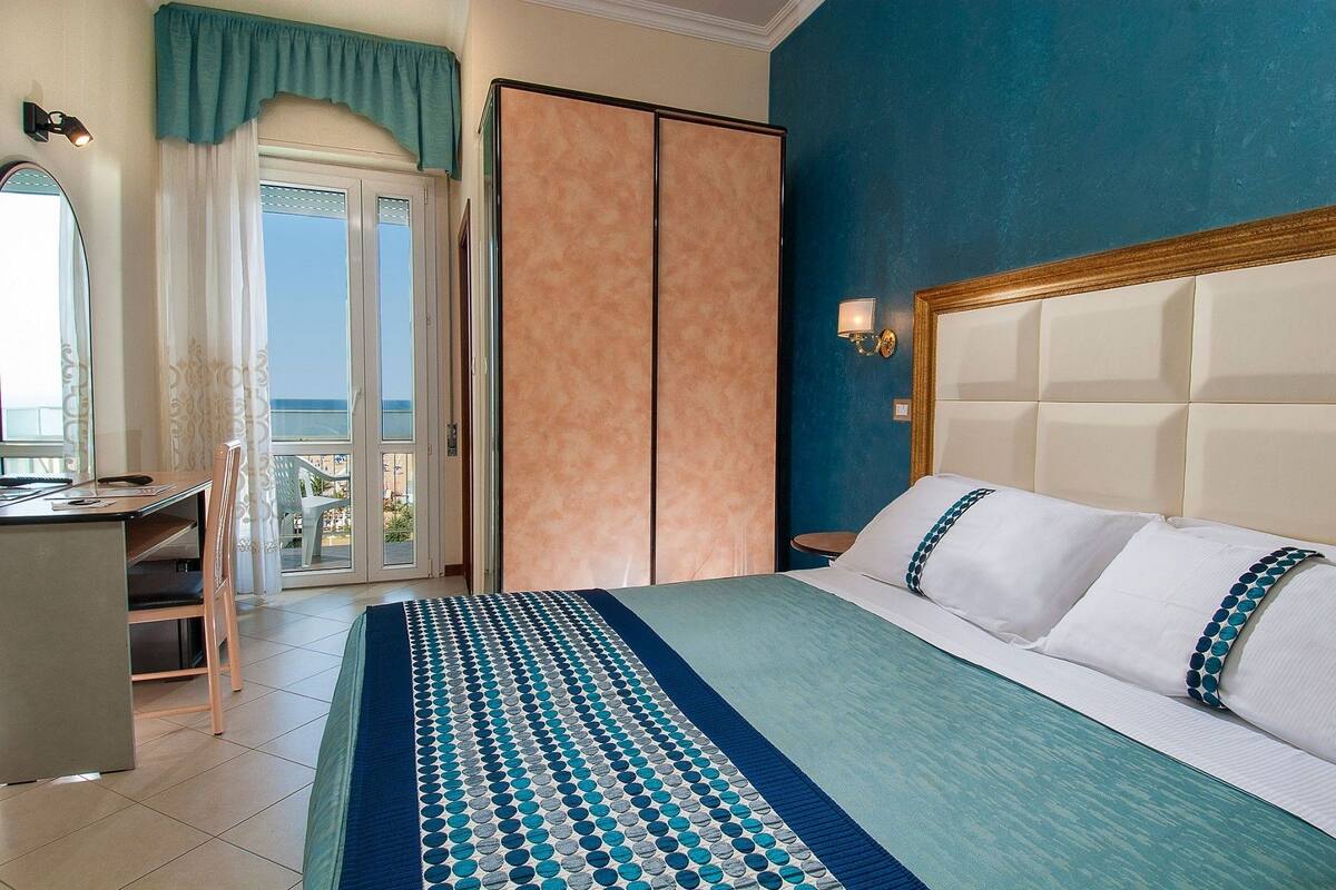 Ein Hotelzimmer mit Bett, Schreibtisch und Meerblick durch eine Glastür.