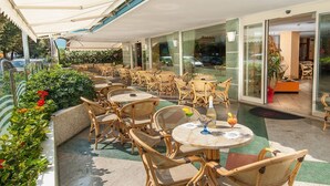 Une terrasse extérieure avec des tables et des chaises en osier, une porte d’entrée en verre et des plantes en pot.
