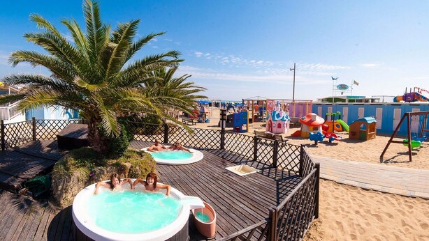 Une terrasse en bois avec des jacuzzis, un palmier et une zone de plage de sable équipée d’aires de jeux pour enfants.