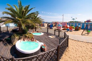 Eine Holzdielen-Terrasse mit Whirlpools, einer Palme und einem Sandstrand mit Spielgeräten für Kinder.