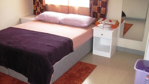 A single bed with a purple blanket, a white nightstand with a phone and a lamp, and a trash bin. - Velvet House (Nairobi)