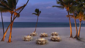 Un espace de restauration aménagé sur la plage, avec des tables et des chaises disposées sur le sable, entouré de palmiers.