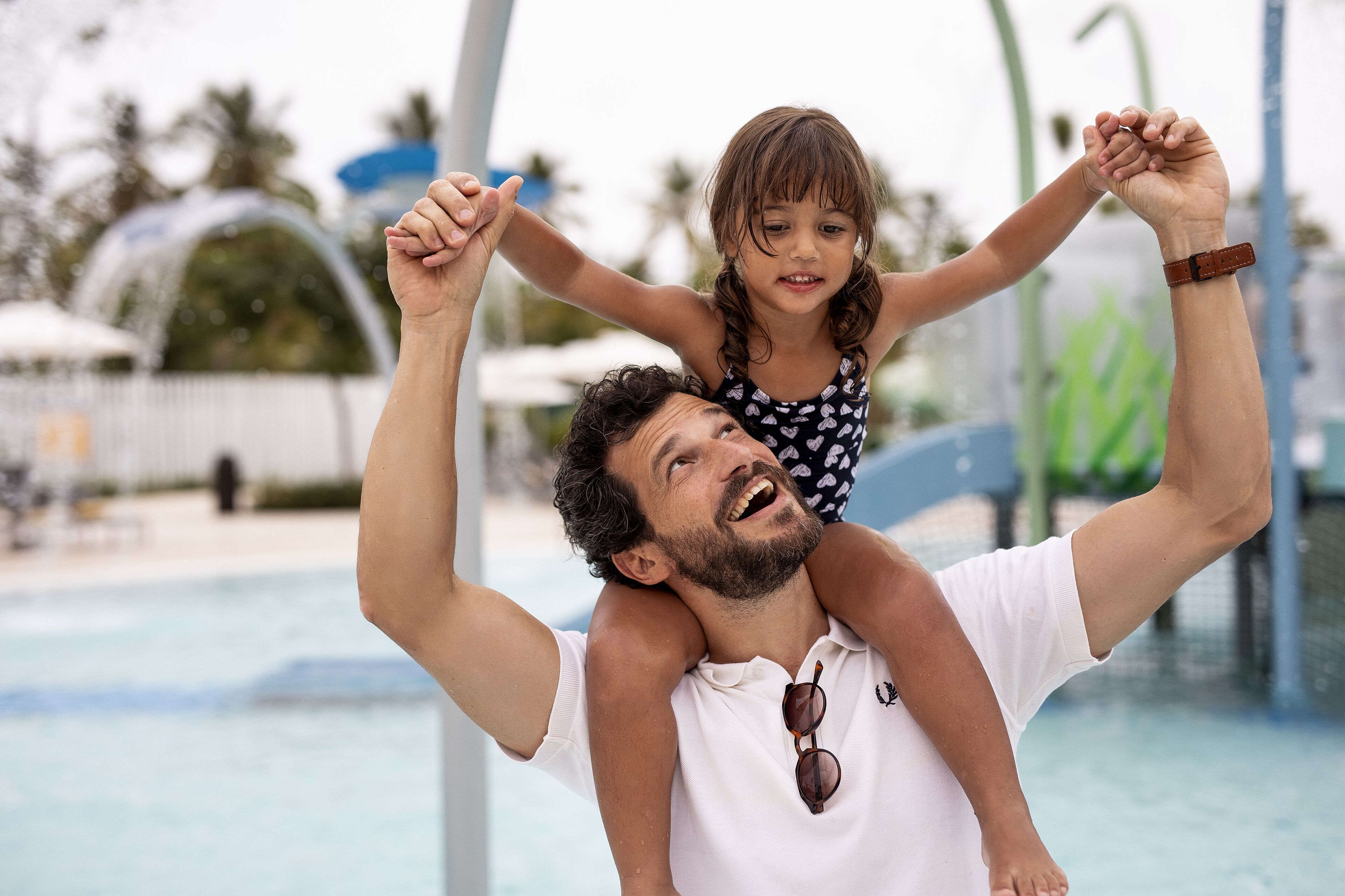 A man carrying a child on his shoulders by a poolside.