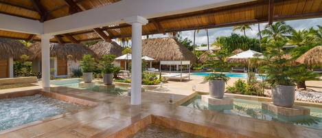 A pool area with multiple hot tubs, thatched-roof structures, and potted plants.