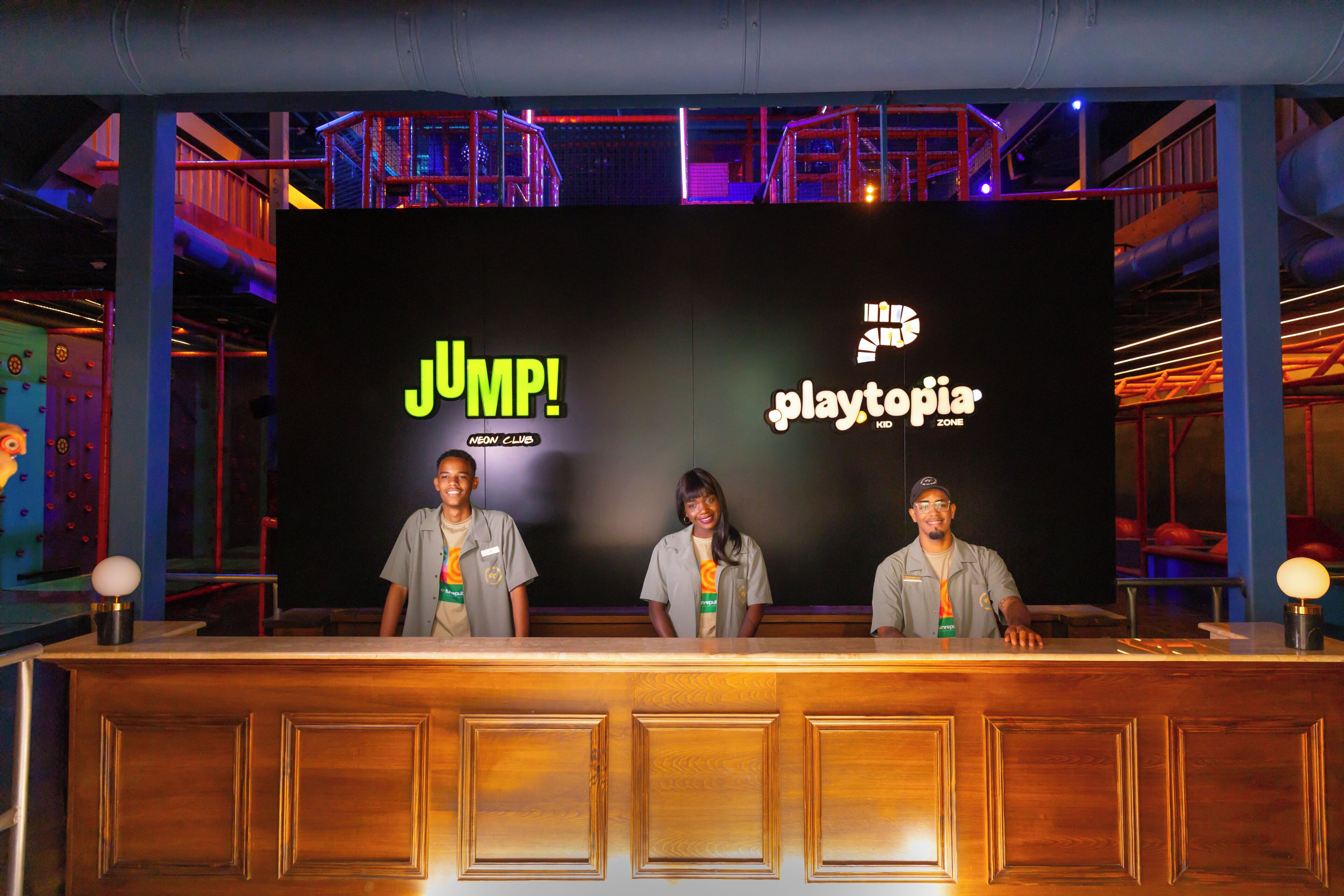 Un mostrador de recepción con un fondo negro que muestra los logotipos 'JUMP!' y 'playtopia', tres empleados con uniforme y un techo de estilo industrial moderno.