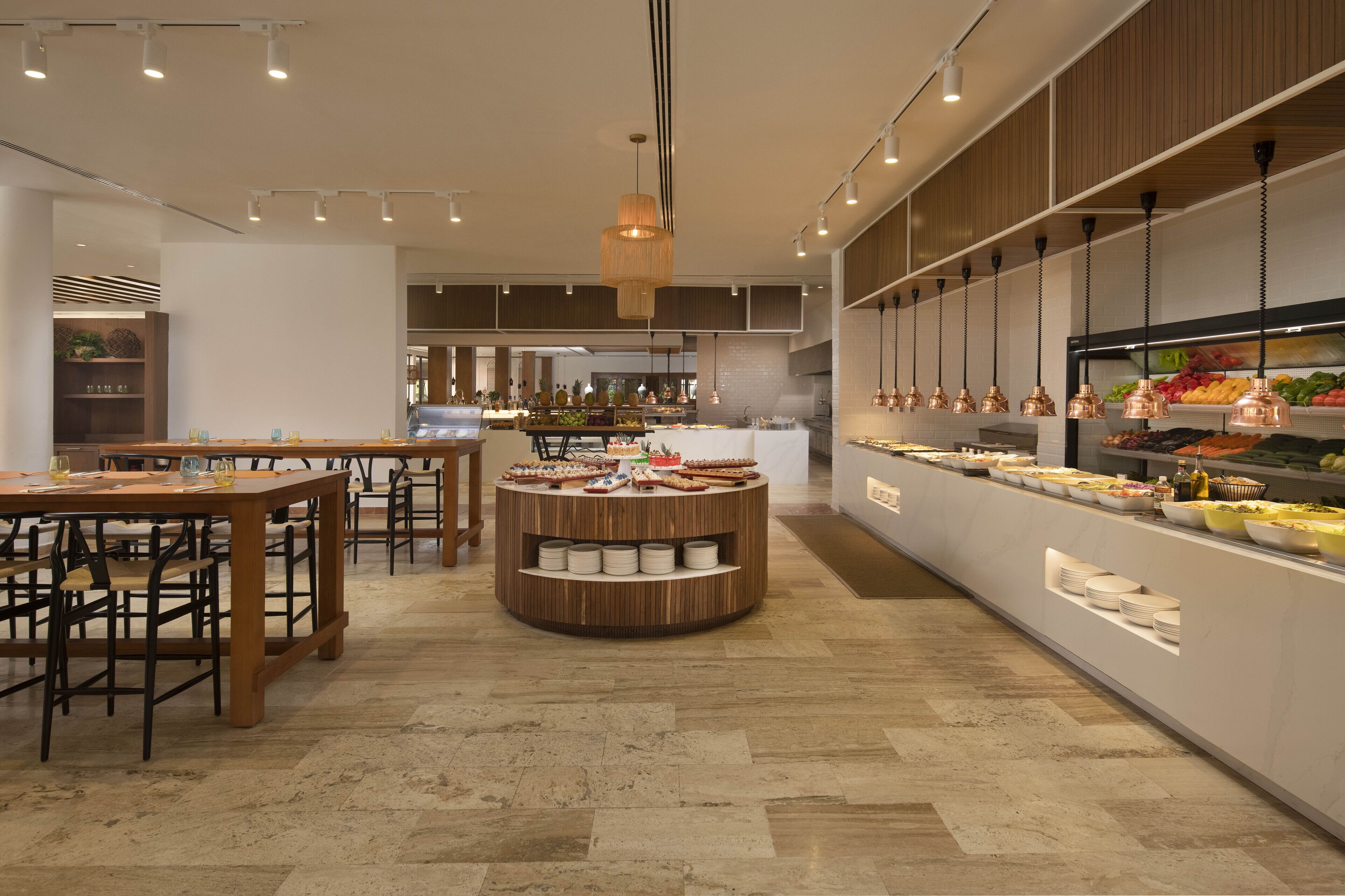 A modern restaurant with a central round table surrounded by chairs, a buffet counter with various food items, and overhead lighting.