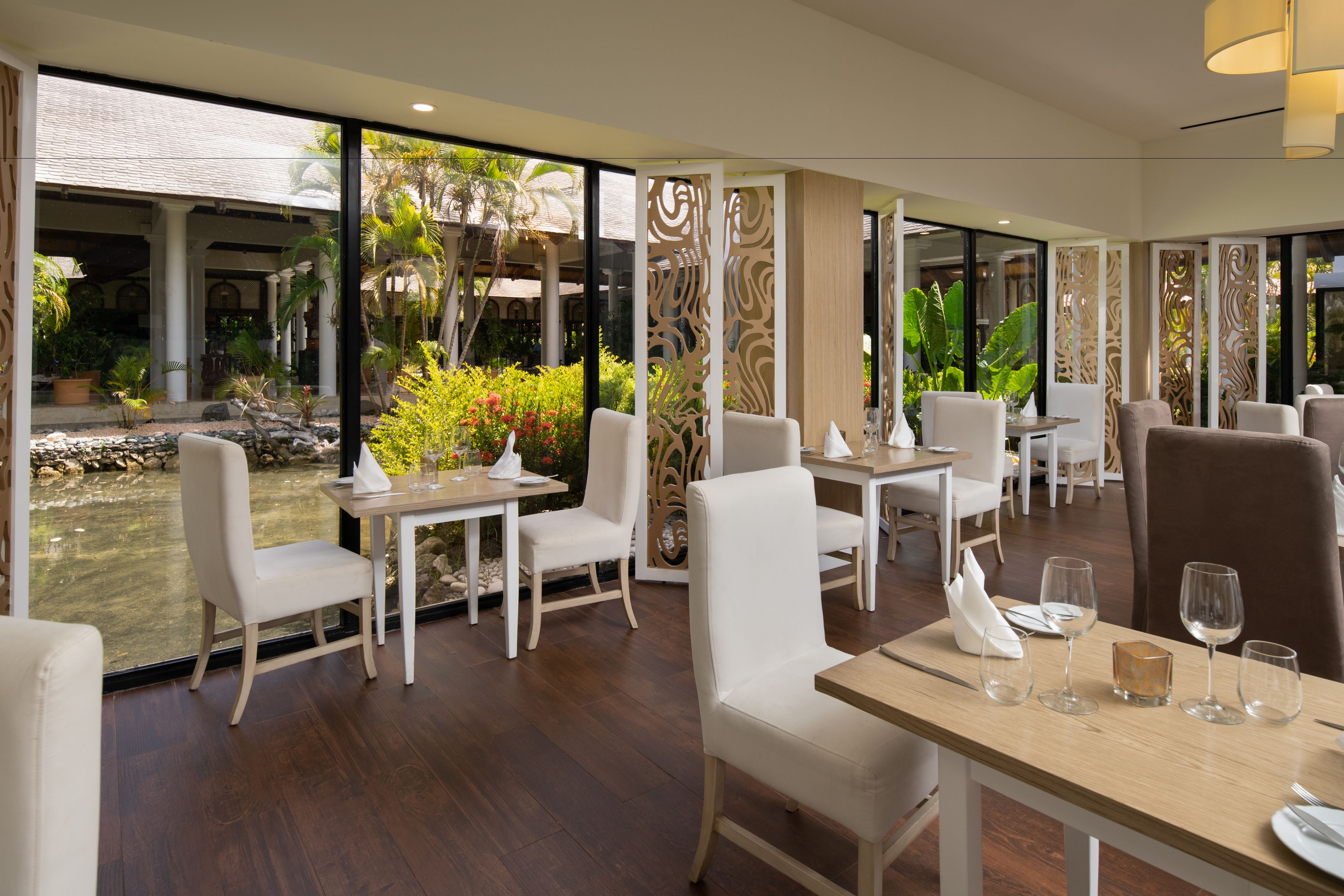Ein Restaurant mit weißen Stühlen und Tischen, mit Blick auf einen Garten mit Palmen und einem Teich.