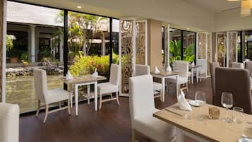 A restaurant with white chairs and tables, a view of a garden with palm trees and a pond.