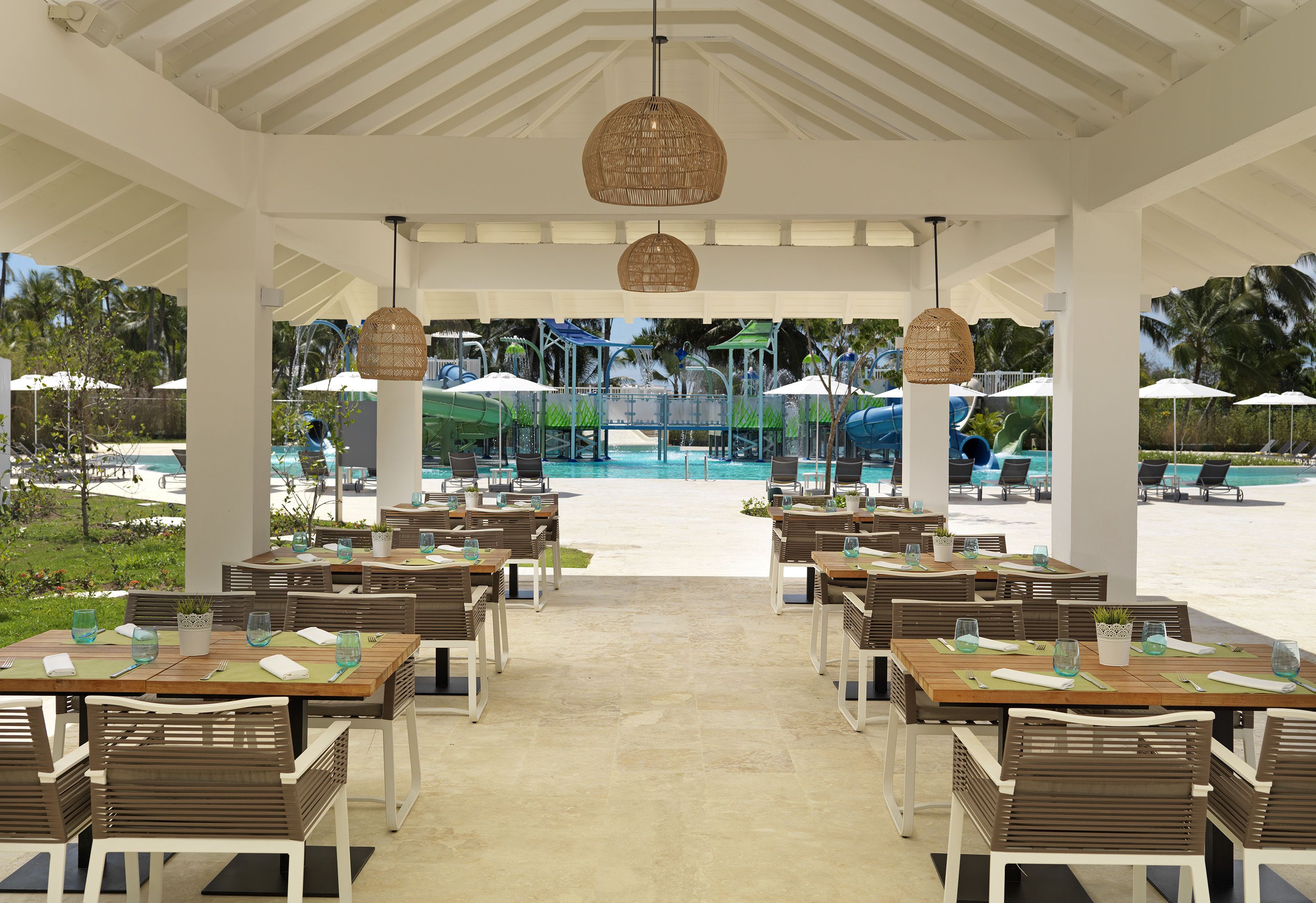 An outdoor dining area with tables and chairs, a pool in the background, and umbrellas.