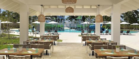 An outdoor dining area with tables and chairs, a pool in the background, and umbrellas.