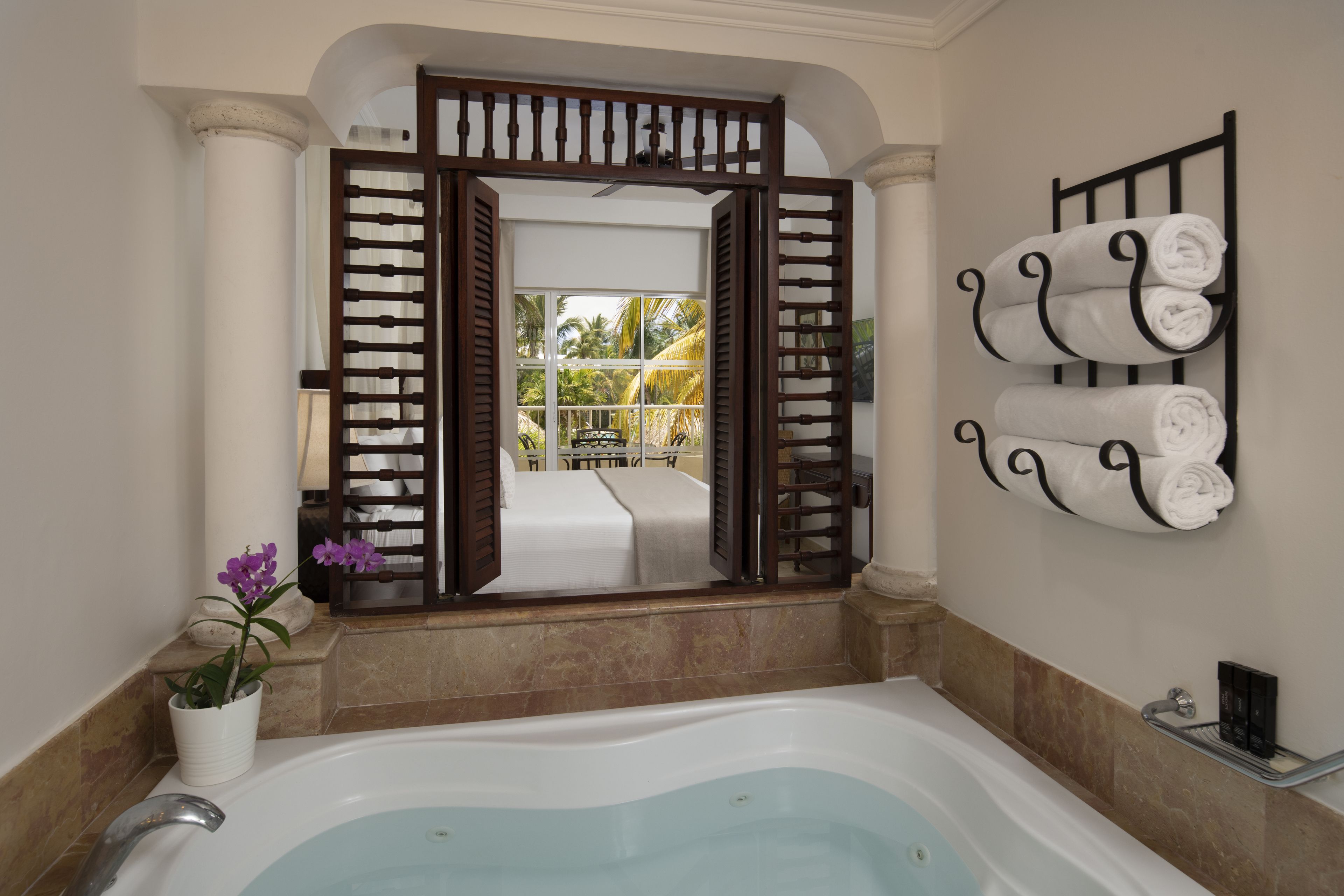 Une salle de bain avec une baignoire, un miroir qui reflète un balcon avec un lit, et un porte-serviettes avec des serviettes.
