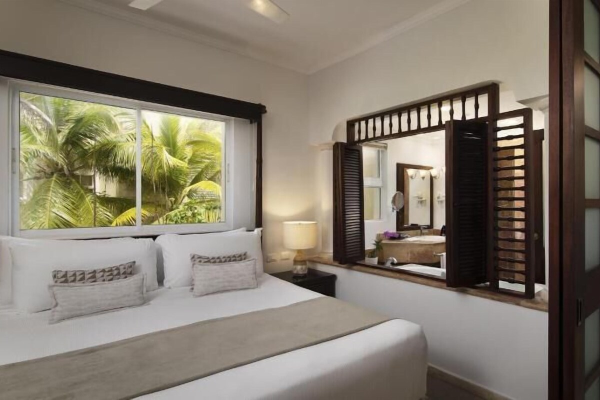 Habitación de hotel con cama, mesita de noche, lámpara, ventana grande con vistas a palmeras y espejo con marco de madera.