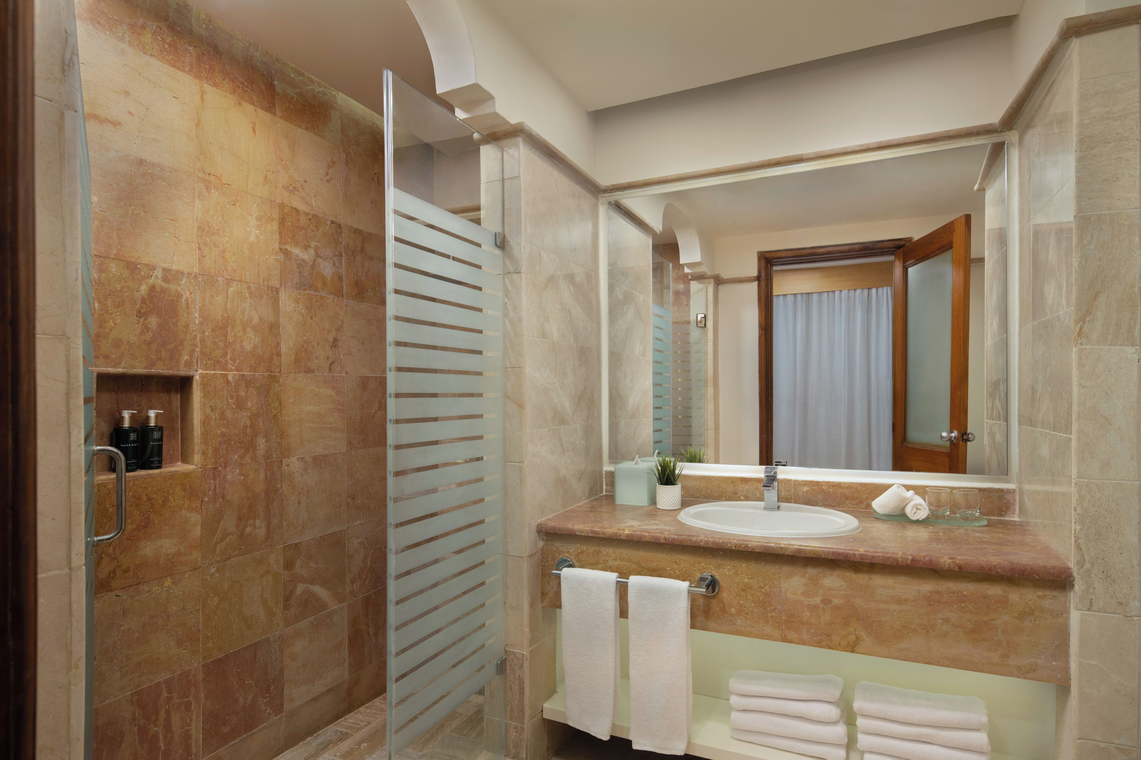 A bathroom with a marble sink, a glass shower enclosure, and a mirror reflecting a door with curtains.
