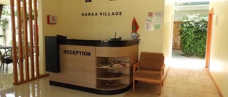 Zona de recepción con mostrador identificado como "RECEPTION", dos relojes de pared y un cartel que dice "KARAA VILLAGE".
