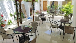 Un espace extérieur aménagé en terrasse, avec des tables rondes et des chaises en osier, entouré de plantes en pot et de palmiers.