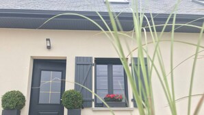 Une maison avec une porte noire, deux fenêtres avec des volets et un petit jardin agrémenté de plantes et de fleurs.