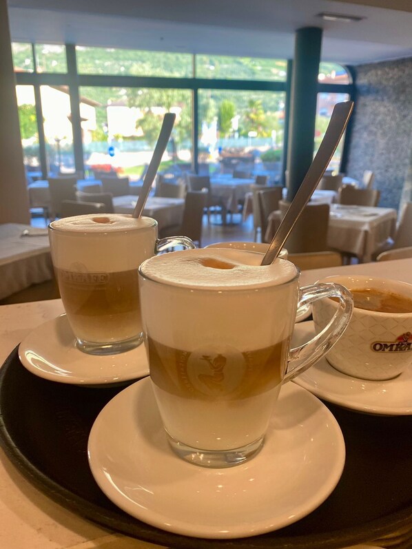 Two cups of coffee with milk on a tray in a restaurant with large windows and outdoor seating visible. - Athos Hotel (Riva del Garda)