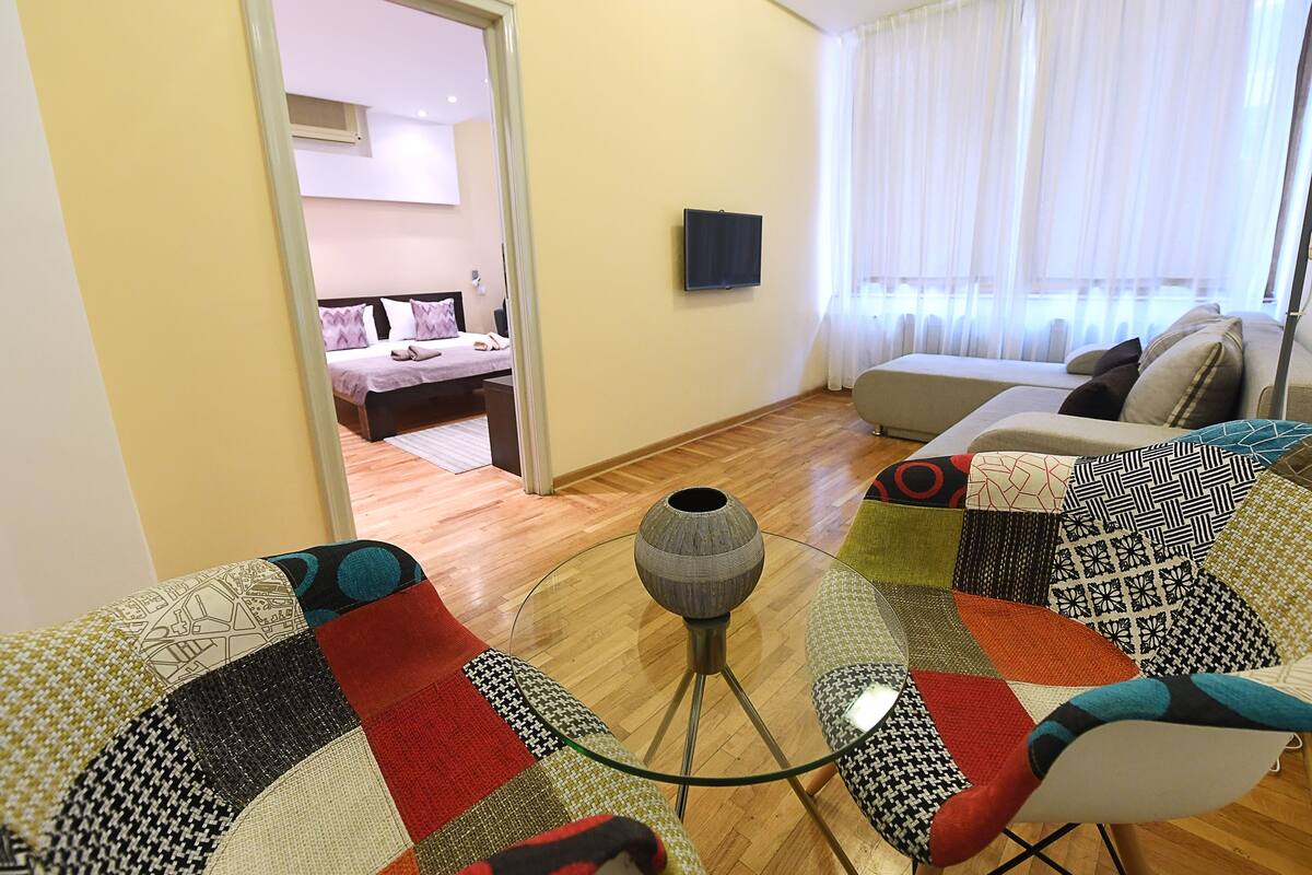 Una sala de estar con una mesa de centro de vidrio, cojines estampados, un sofá y una cama al fondo.