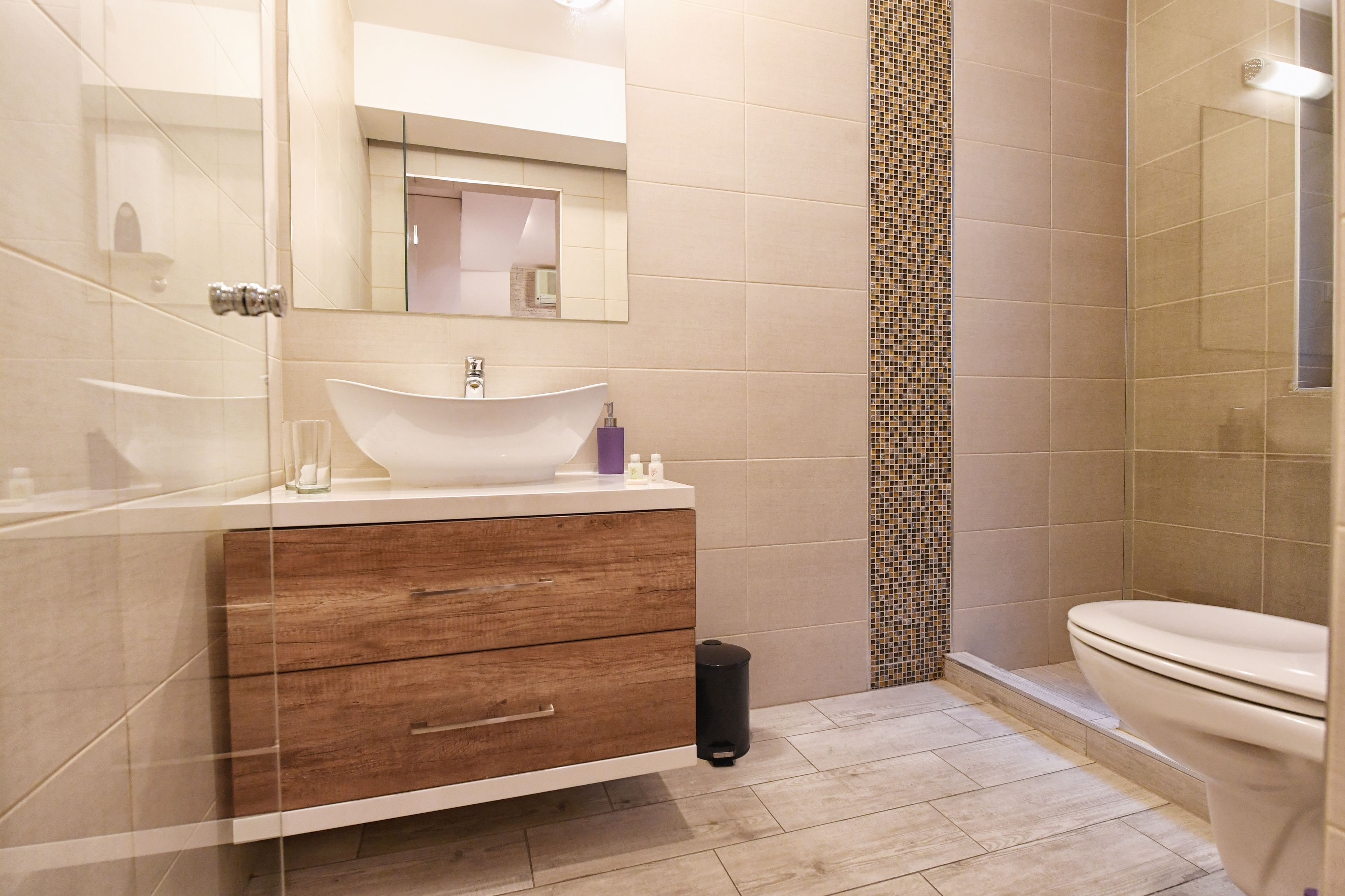 A modern bathroom with a large mirror, a white sink, wooden vanity, and a tiled shower area.