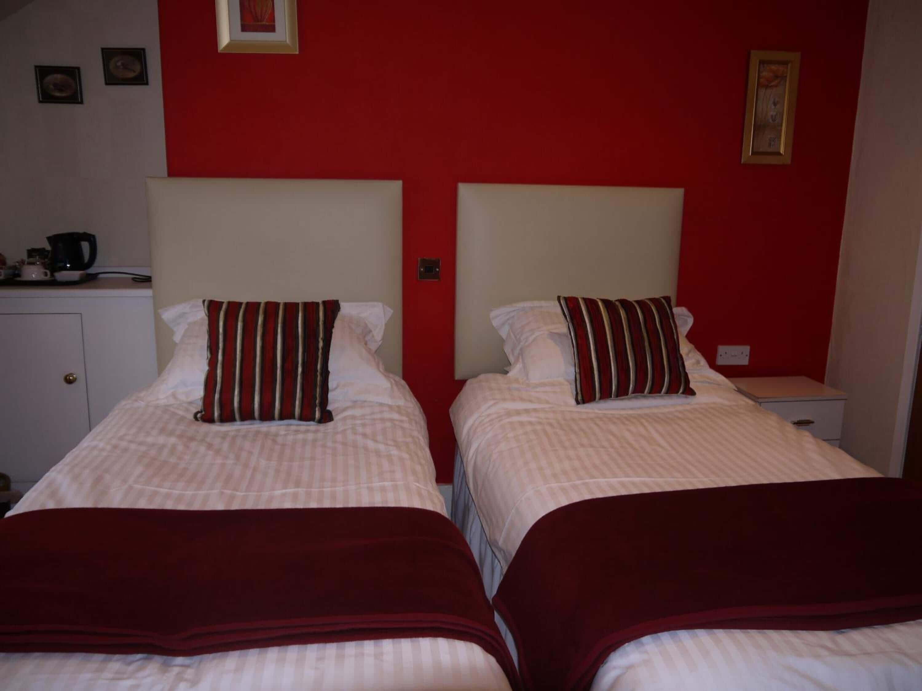Dos camas individuales con ropa de cama a rayas y almohadas rojas, una mesita de noche pequeña y una pared roja con cuadros enmarcados.