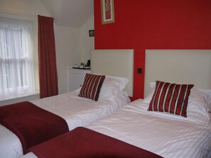 Habitación de hotel con dos camas, cortinas rojas, un cabecero blanco y una pequeña mesa con cafetera.