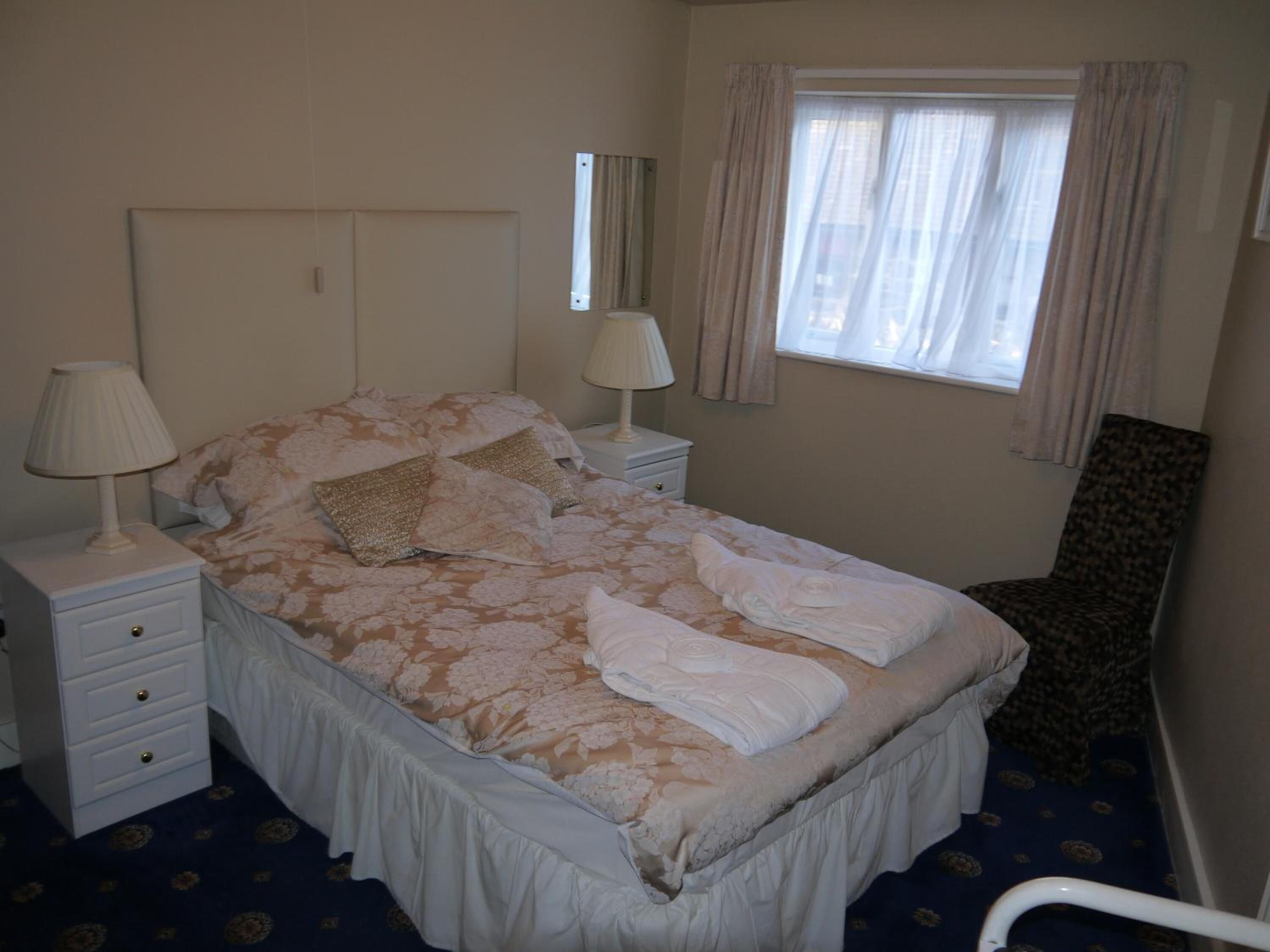 A neatly made bed with floral bedding, two bedside lamps, a chair, and a window with curtains.