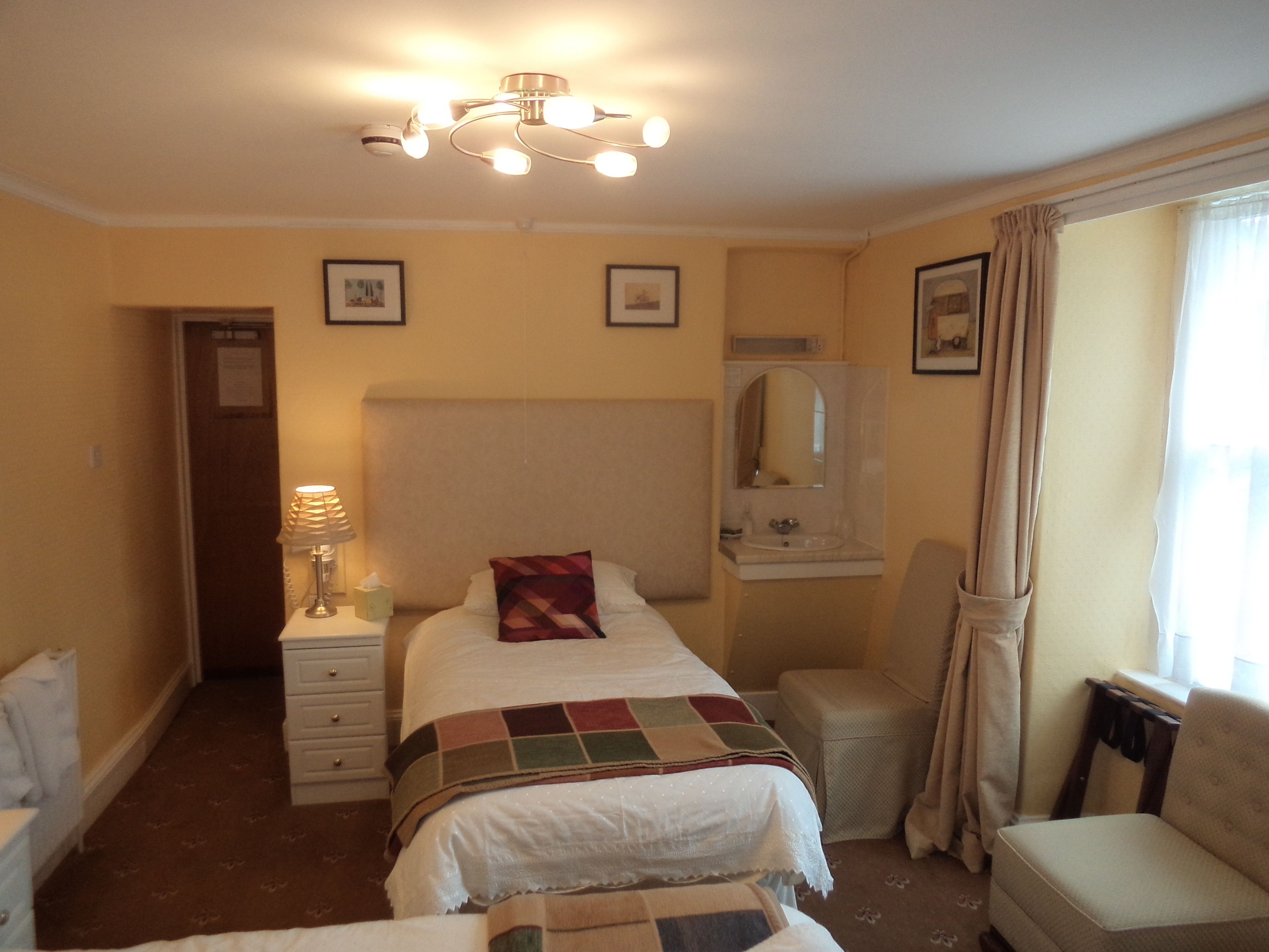 A hotel room with a bed, a bedside table with a lamp, a chair, a window with curtains, and a mirror above the sink.