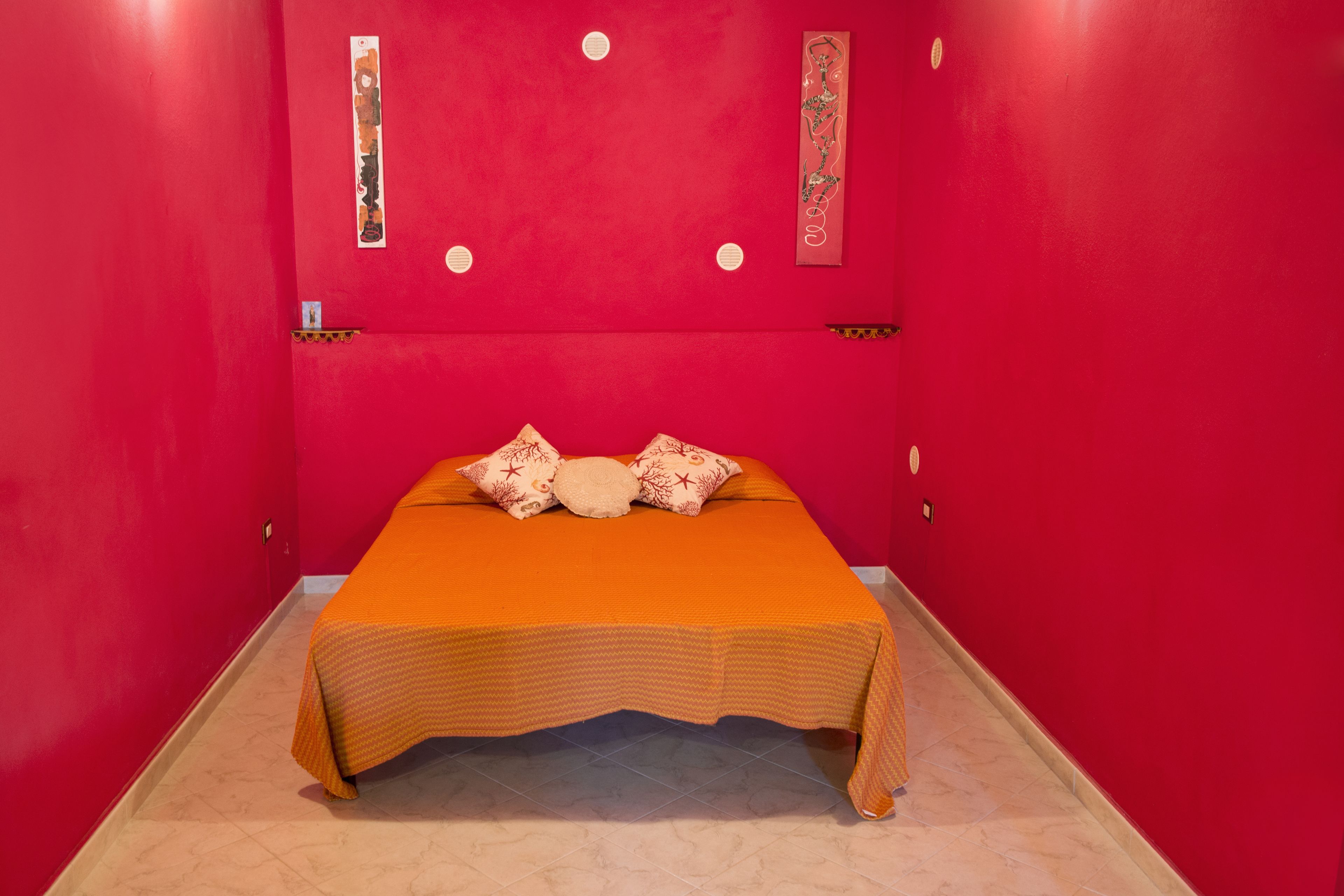 Una habitación pequeña y de colores vivos, con una cama individual, una pared roja y un estante con adornos.