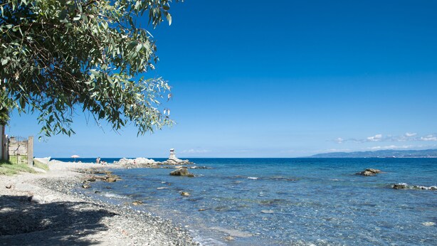 A beach with clear water, pebbles, a lighthouse, and a building on the shore.