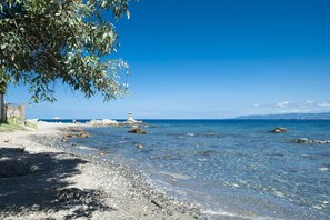 A beach with clear water, pebbles, a lighthouse, and a building on the shore. - Sabrina Suite (Milazzo)
