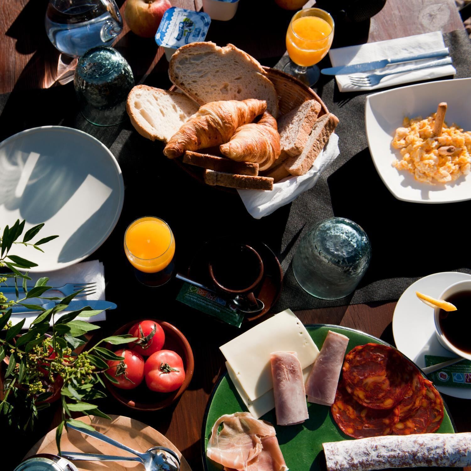 A breakfast table with croissants, bread, tomatoes, cheese, ham, and various condiments.
