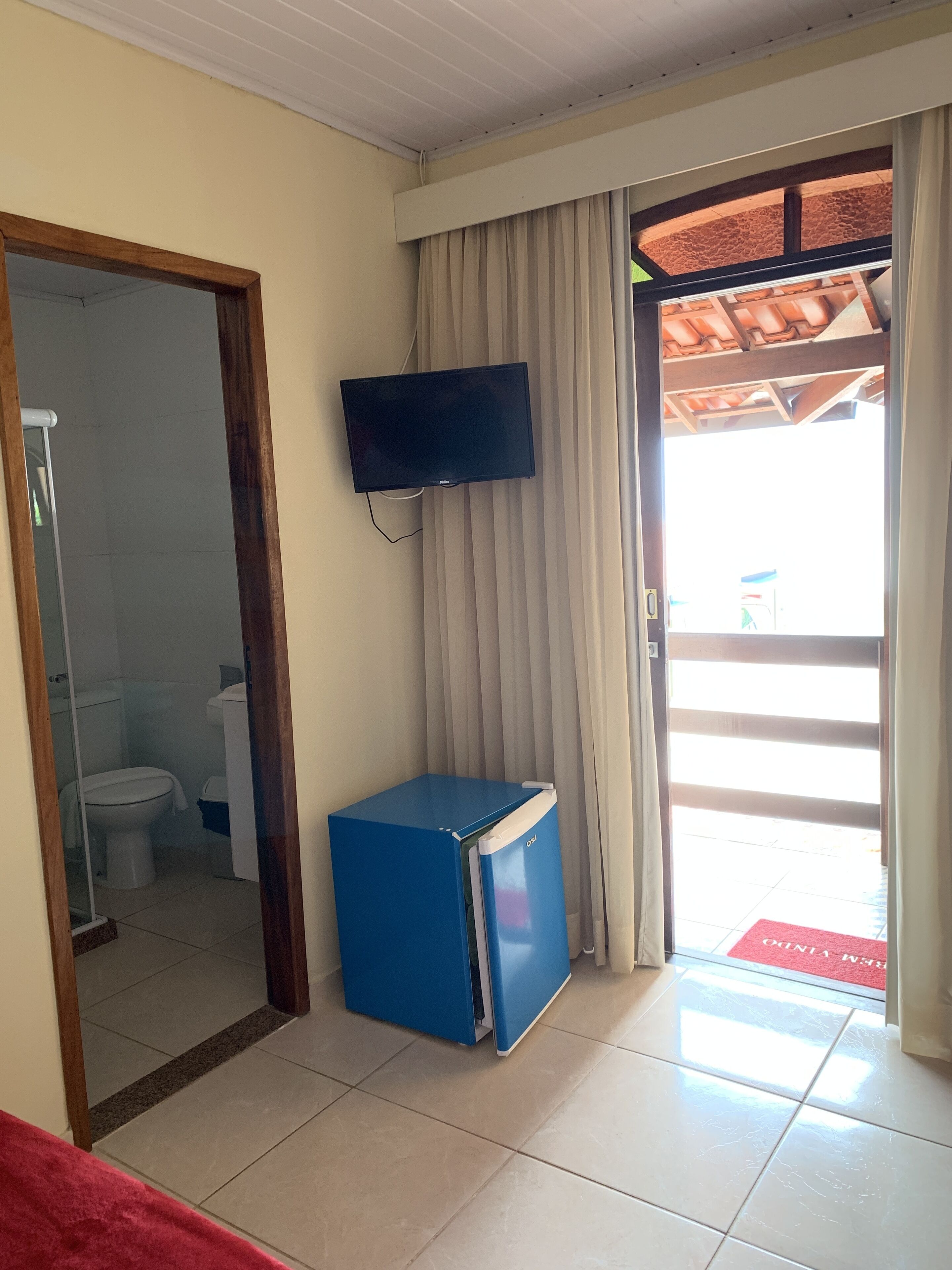 A room with a television mounted on the wall, a blue refrigerator, a bathroom with a toilet and sink, and a door leading outside.
