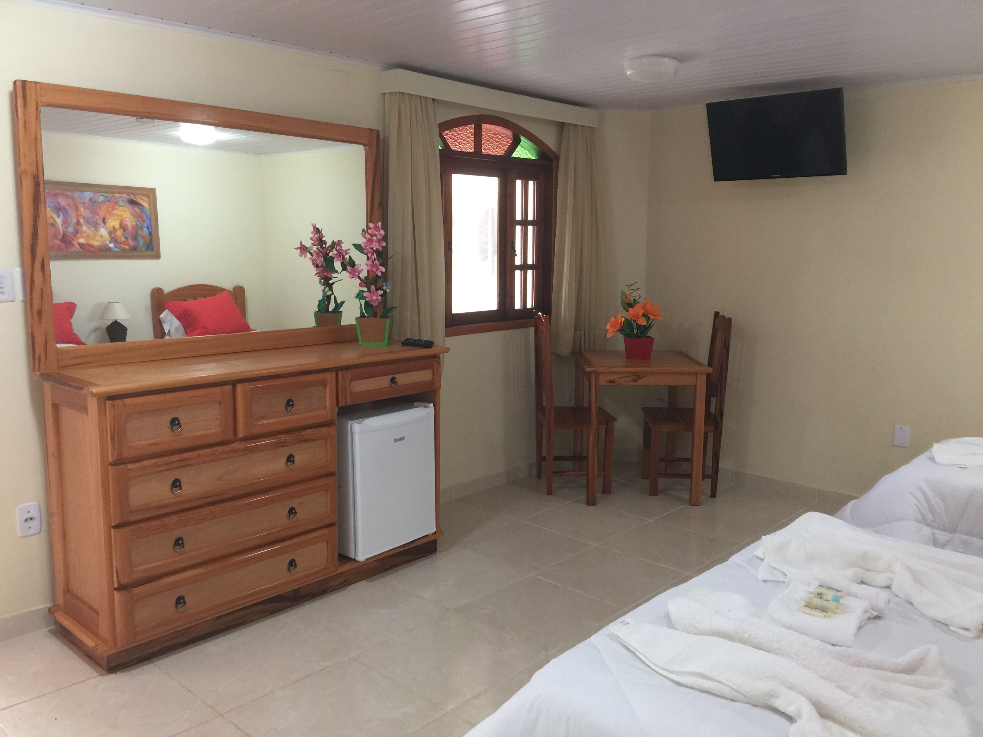 Une chambre d’hôtel avec une commode en bois, un petit réfrigérateur, un lit avec du linge de maison blanc, une table à manger avec des chaises et un grand miroir.