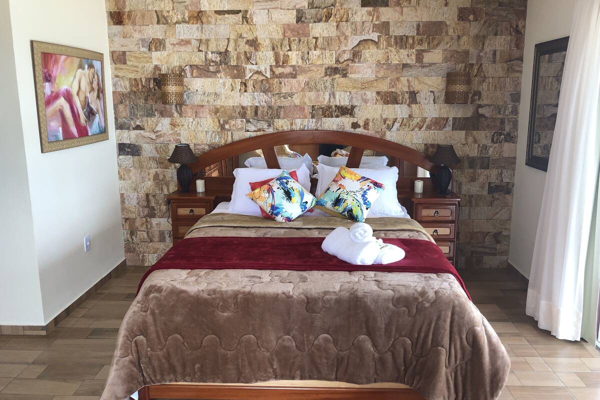 Cama bem arrumada com cobertura marrom, cabeceira de madeira e várias almofadas.