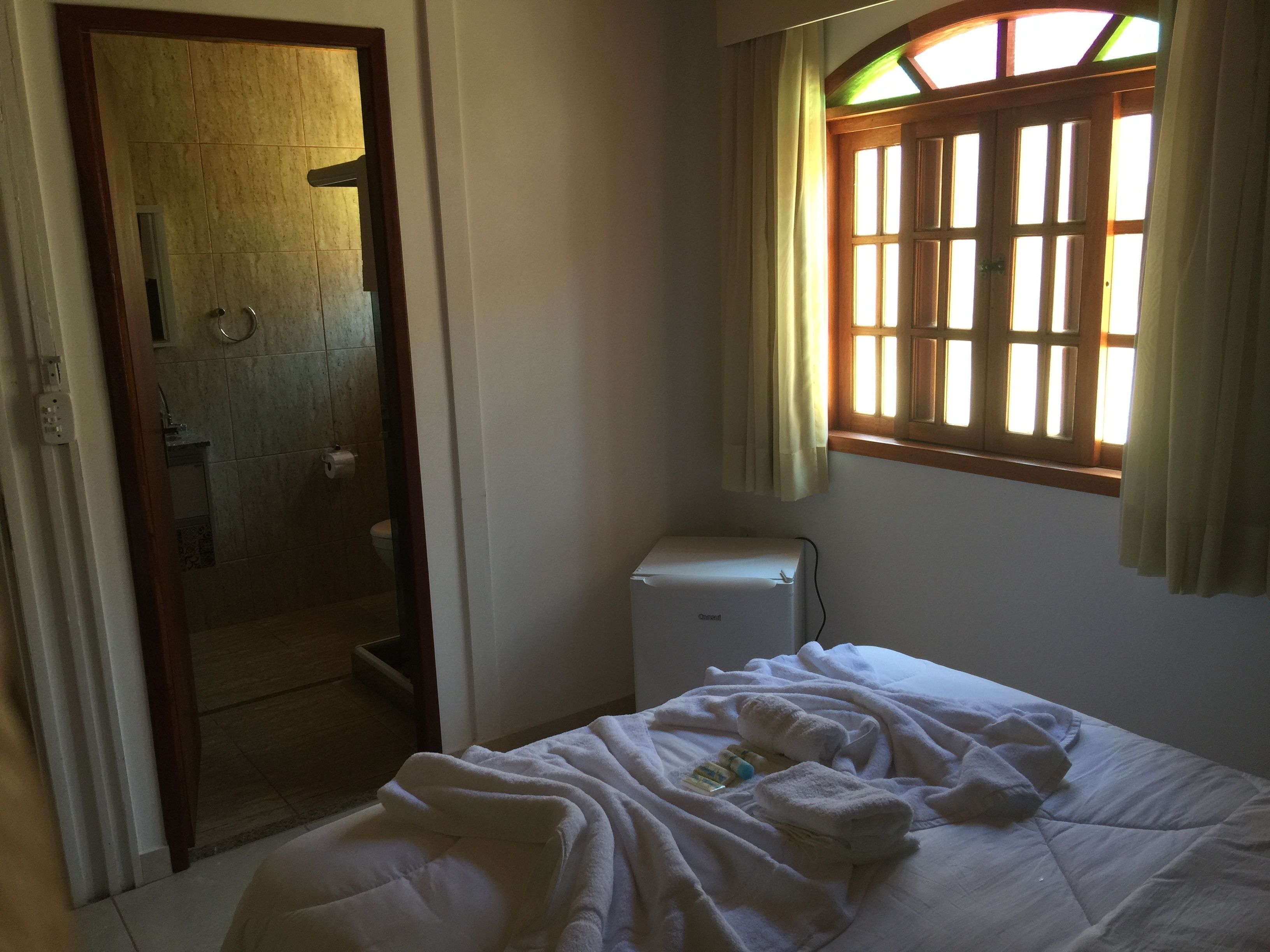 Un dormitorio con cama, ventana con cortinas, radiador y puerta que da a un baño.