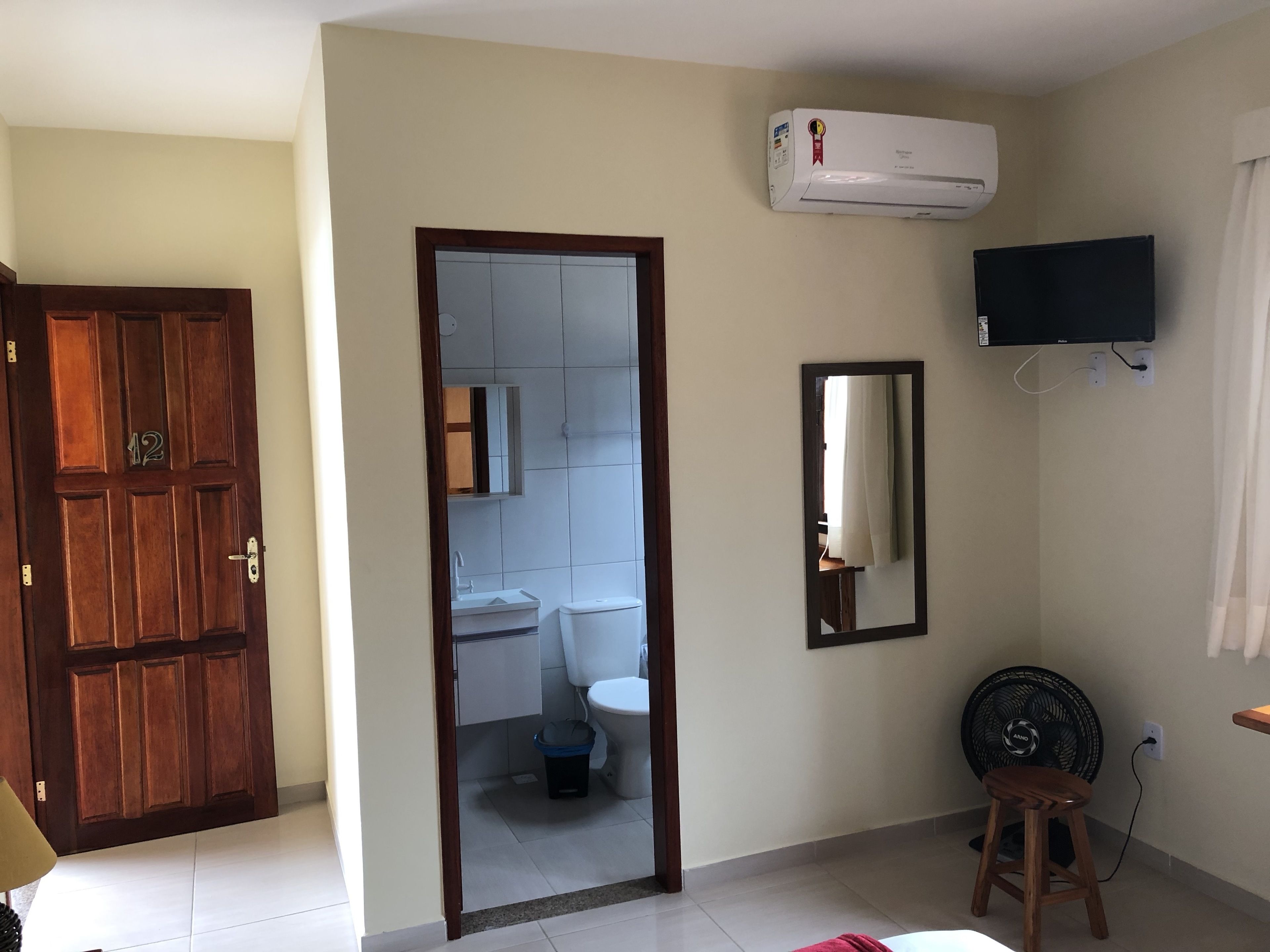 Una habitación con puerta de madera, un espejo, un abanico negro y un equipo de aire acondicionado en la pared.
