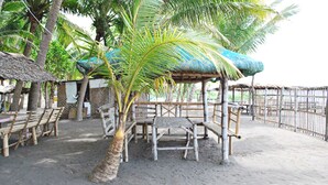 Un área de estar frente a la playa con sombrillas de paja, muebles de madera y palmeras.