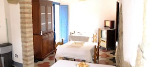 A small, cozy room with a bed, a table with a basket of bread, a wooden cabinet, a blue door, and a television.