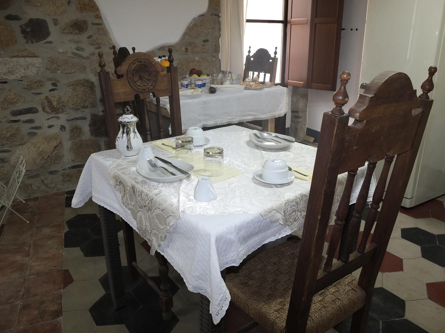 Una mesa de comedor con mantel blanco y cubertería, rodeada de paredes de piedra y una silla de madera con cojín de asiento tejido.